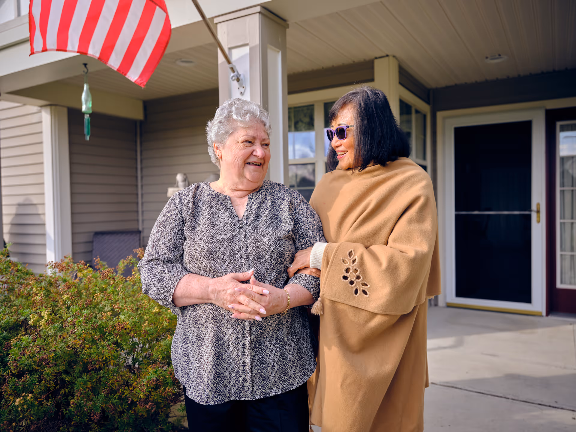 Two women standing outside a building entrance, smiling and holding arms. One woman has gray hair and is wearing a patterned blouse, while the other woman has dark hair, sunglasses, and a tan shawl. An American flag is visible above them, and there are bushes and a porch in the background.