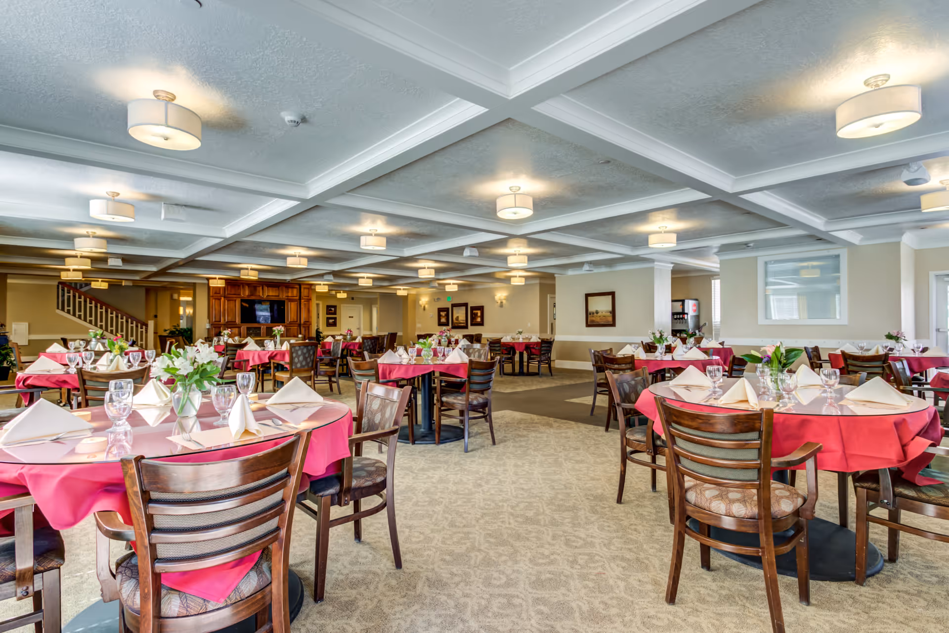 A spacious senior living dining room with multiple round tables covered in red tablecloths and set with folded napkins, glassware, and small floral centerpieces. The room features a coffered ceiling with multiple light fixtures, beige walls, carpeted floors, and wooden chairs. In the background, there is a wooden cabinet with a television and a staircase leading to another level.