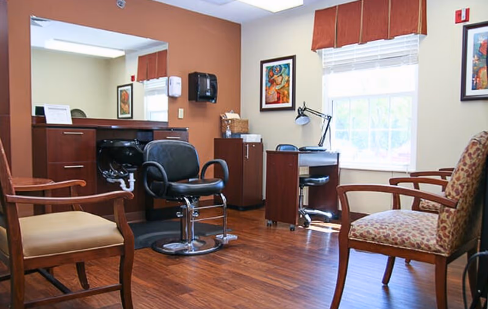 A well-lit room with wooden flooring featuring a black salon chair in front of a large mirror and a sink, a small desk with a lamp and chair near a window with blinds and a red valance, and several wooden chairs with cushioned seats arranged around the room. The walls are painted in beige and brown tones with framed artwork hanging.