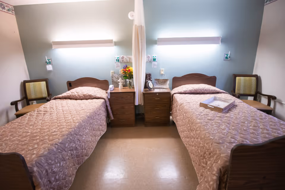 A shared patient bedroom with two single beds, nightstands, chairs, and overhead wall lights.