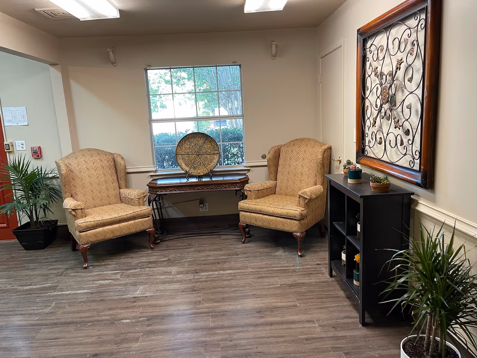 A cozy sitting area with two patterned armchairs facing each other, a wooden table with a decorative plate in front of a window, a black shelving unit with small potted plants, and a large decorative wrought iron wall art piece. There are also two potted plants on the floor and wood-style flooring.
