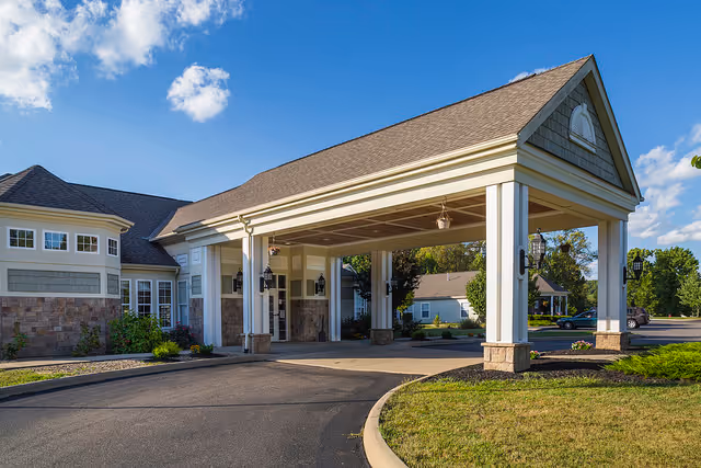 Exterior view of a senior living facility entrance with a covered drop-off area supported by white columns. The building features a combination of stone and siding with multiple windows, surrounded by landscaped greenery and a clear blue sky with a few clouds.