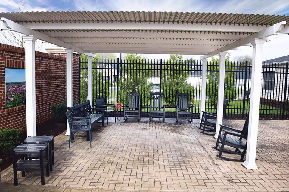 Outdoor patio area with a white pergola overhead, black rocking chairs and benches arranged on a brick-paved floor, a black metal fence in the background, and a brick wall with a framed picture of a flower field on the left side.