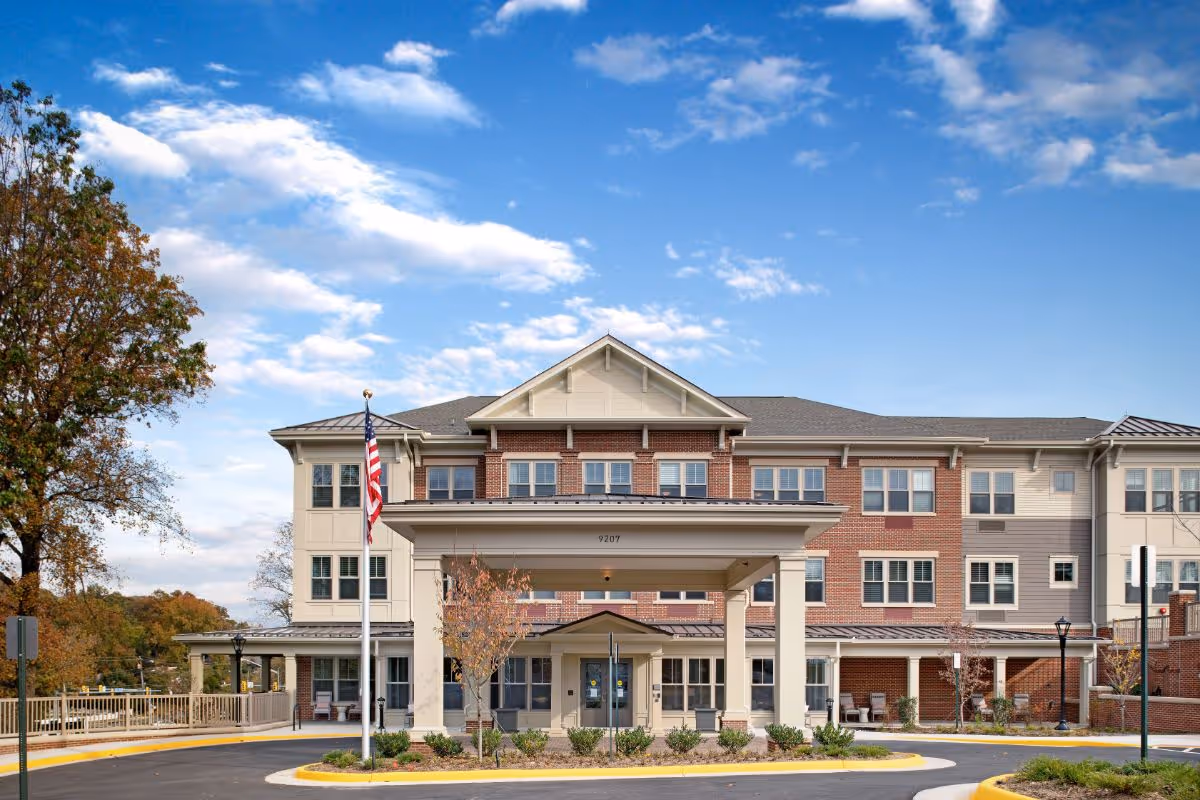 Front exterior view of a three-story senior living facility building with a covered entrance, an American flag on a flagpole, and landscaped greenery under a partly cloudy blue sky.
