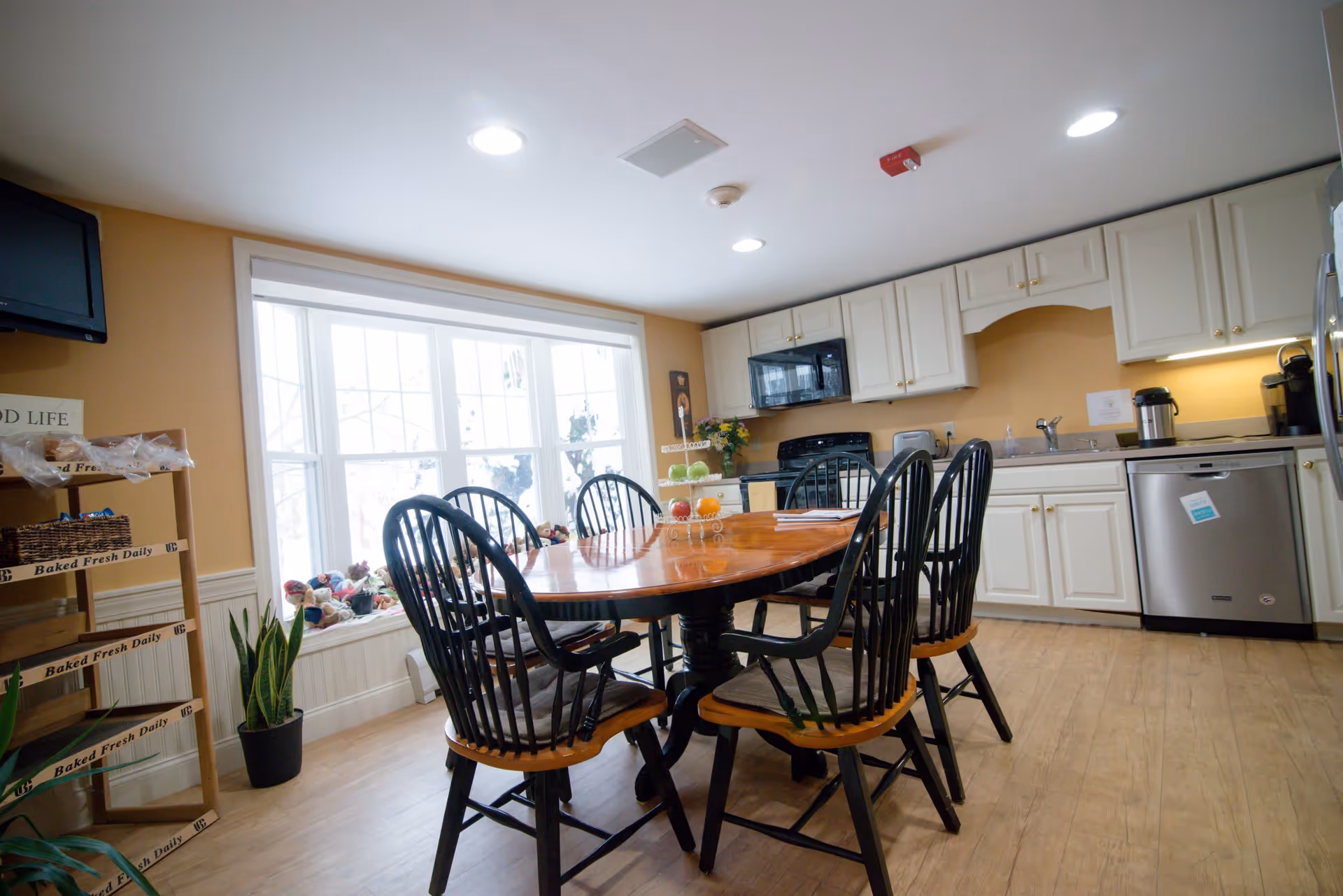 A bright kitchen and dining area with a wooden table surrounded by six black chairs. The kitchen features white cabinets, a black microwave, a black stove, a dishwasher, and a coffee maker. A large window lets in natural light, and there is a wooden rack labeled 'Baked Fresh Daily' with baked goods on the left side. The floor is light wood, and the walls are painted a warm beige color.