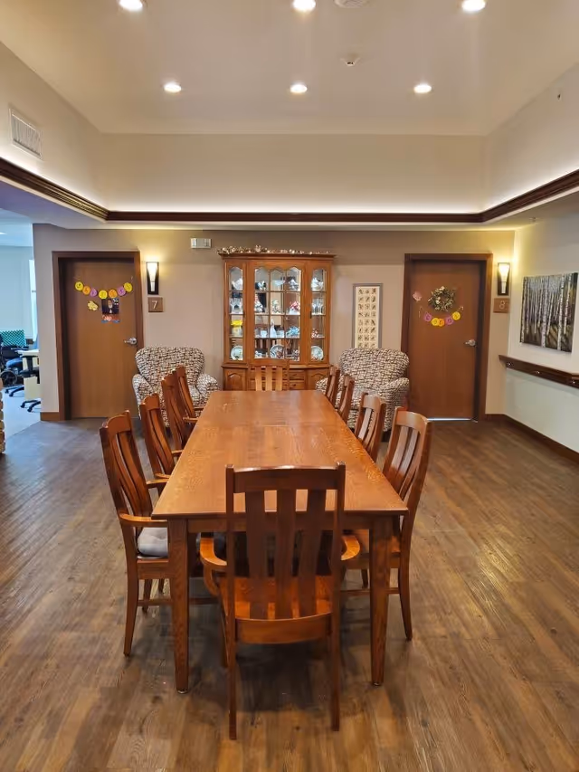 Long wooden dining table with chairs in a communal room, flanked by armchairs and a glass-front china cabinet against the back wall.