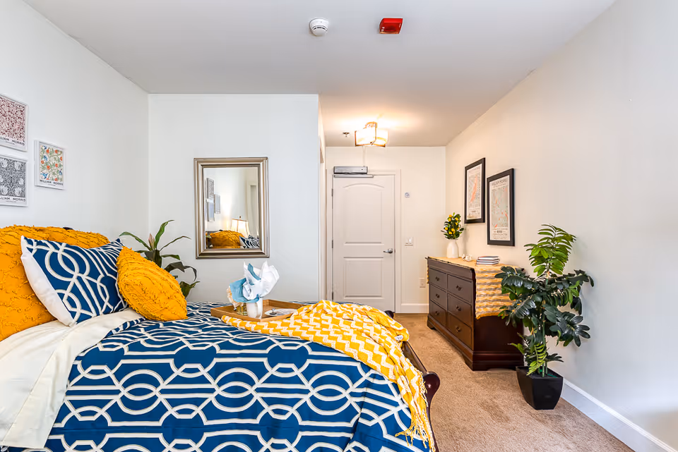 A bright and neatly decorated bedroom featuring a bed with blue and white patterned bedding and yellow accent pillows and throw. There is a wooden dresser with a yellow runner, a vase with flowers, and framed artwork on the wall. A large mirror hangs on the wall near the bed, and a potted plant is placed on the floor. The room has beige carpet and white walls with a closed white door at the far end.