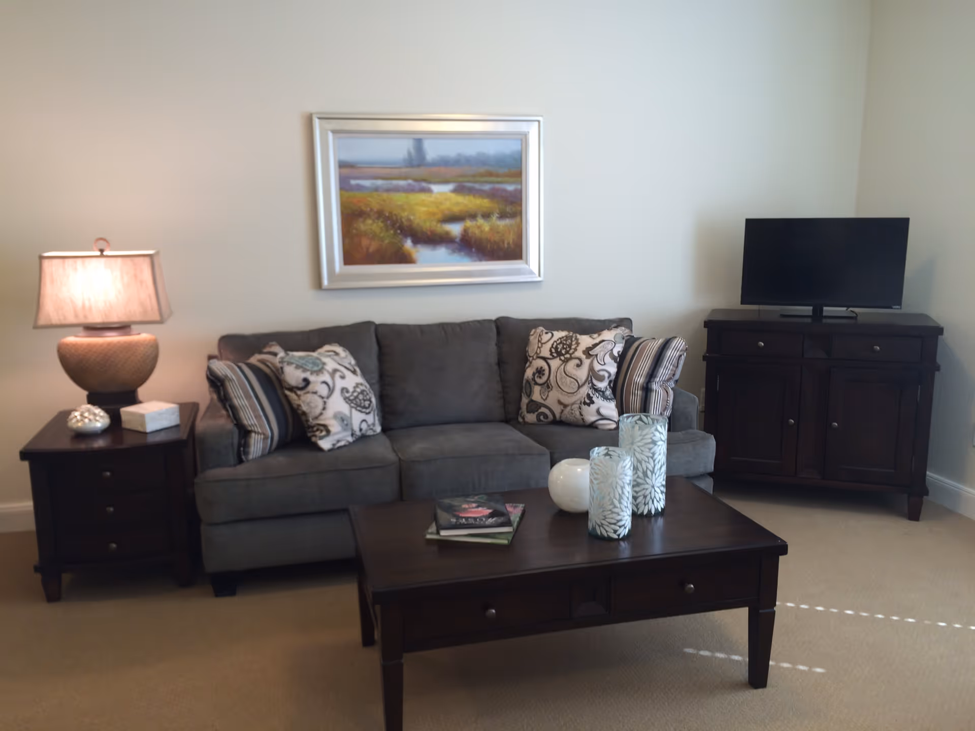 A cozy living room with a gray sofa adorned with patterned cushions, a dark wooden coffee table with decorative items and books, a side table with a lamp, a framed landscape painting on the wall, and a dark wooden TV stand with a flat-screen television.