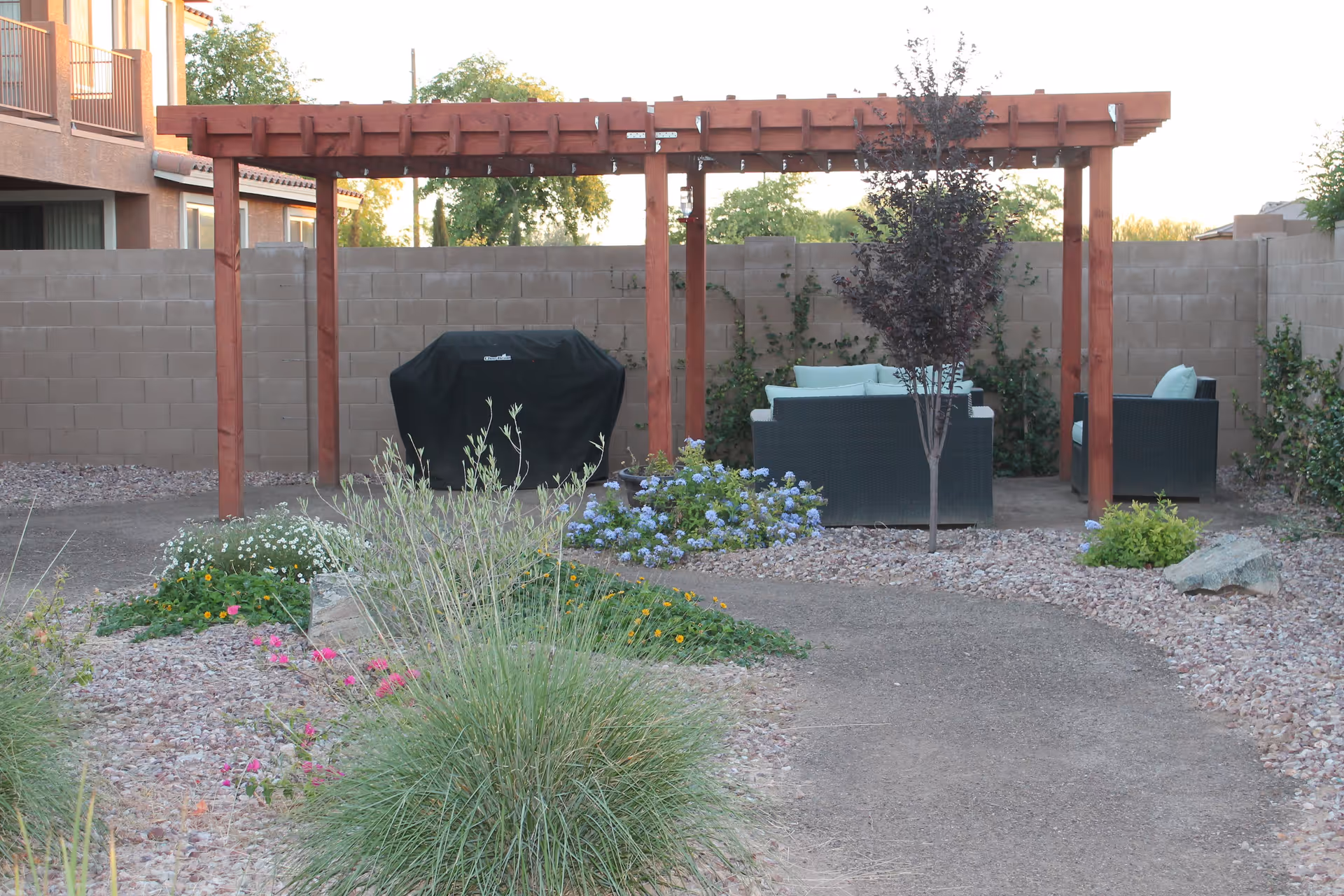 Outdoor patio area with a wooden pergola, a covered barbecue grill, cushioned wicker chairs, a small tree, and various plants and flowers surrounded by a gravel pathway and a block wall fence.
