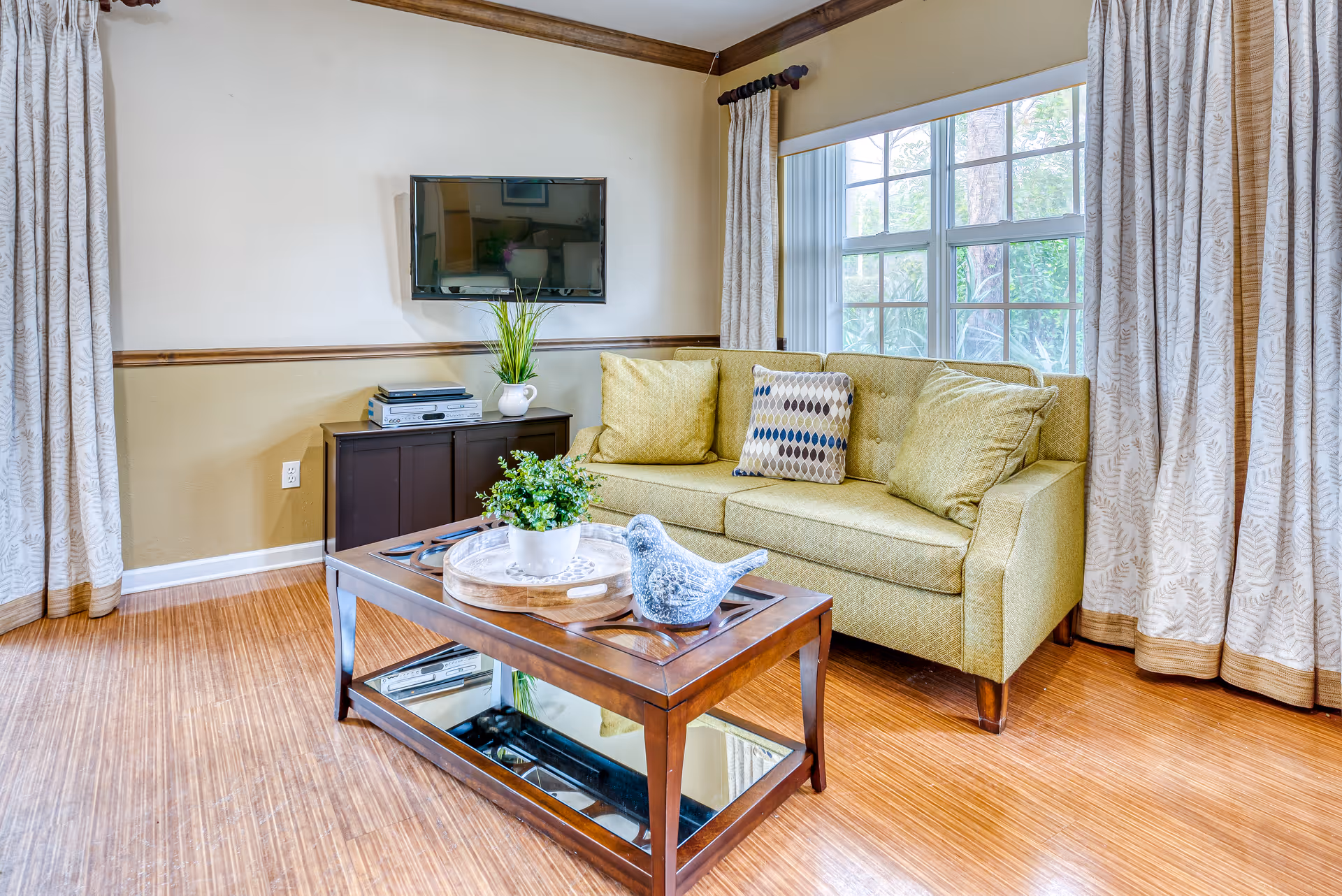 Bright living room with a green loveseat, coffee table with decorative items, wall-mounted TV, and a large window with curtains.