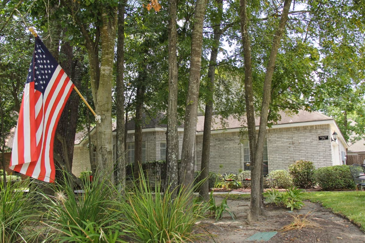 Exterior view of a single-story brick building partially obscured by several trees and greenery. An American flag is mounted on a pole attached to one of the trees in the foreground. The building has a sign that reads 'Cottage #4'.