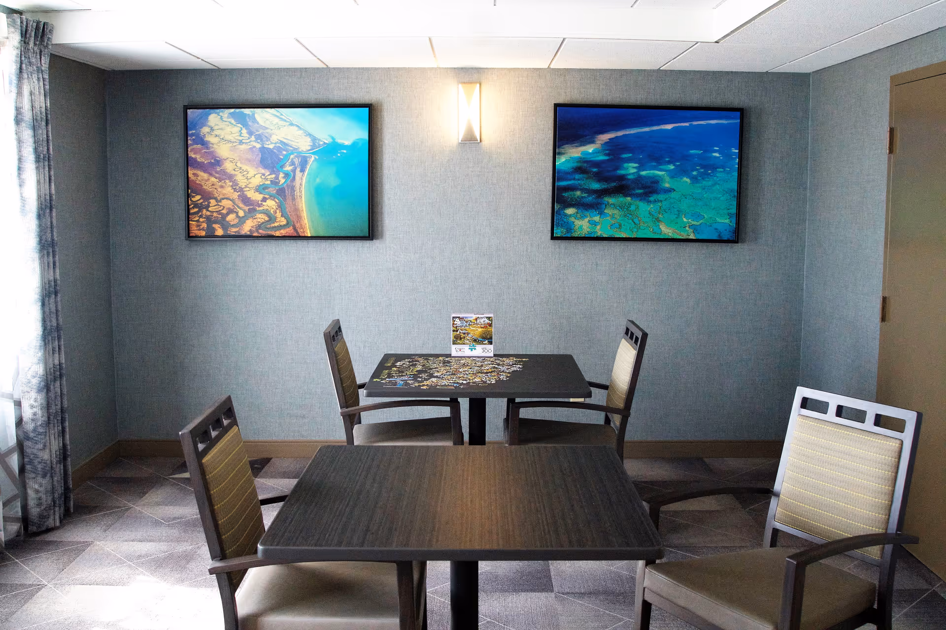 Small common room with two square tables and four chairs, a jigsaw puzzle on one table, and two framed ocean photographs on the wall.