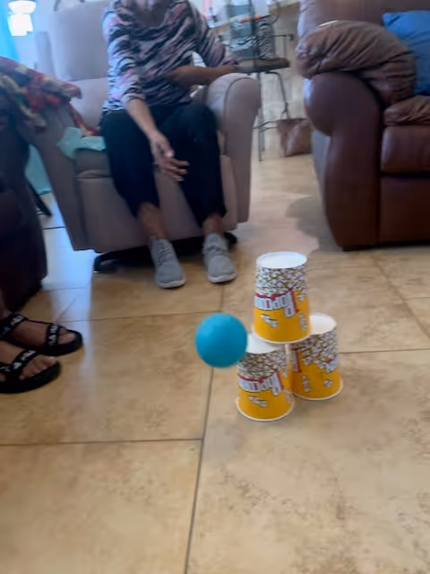 Seniors sitting in a living room area with three stacked popcorn cups and a blue ball on the tiled floor.
