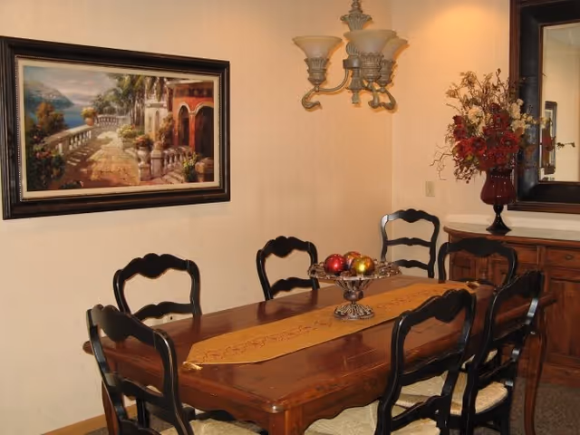 A dining room with a wooden table and six black chairs. The table has a decorative runner and a centerpiece bowl with colorful decorative balls. On the wall is a framed painting of a scenic outdoor terrace with palm trees and a body of water. A wall-mounted light fixture with three lamps is above the table. To the right, there is a wooden sideboard with a large mirror above it and a vase with red and white flowers.