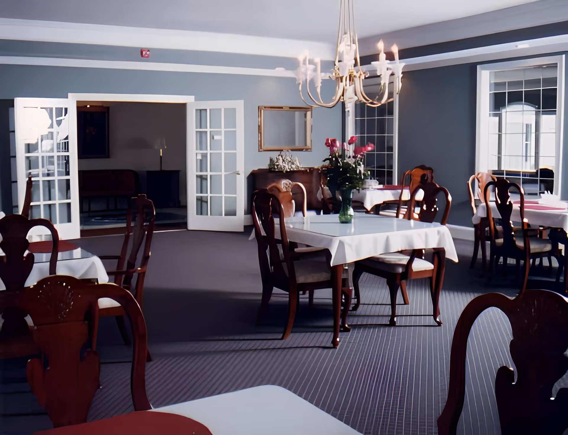 A bright formal dining room with tables draped in white tablecloths, wooden chairs, a chandelier, and French doors opening to another room.