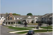 Front exterior view of a large senior living facility building with multiple windows, a covered entrance, and a circular driveway with parked cars. The building is surrounded by well-maintained lawns and small trees.