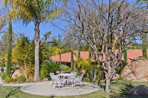 Outdoor seating area with white metal table and chairs on a circular concrete patio surrounded by palm trees, other greenery, and a building with a red-tiled roof in the background under a clear blue sky.