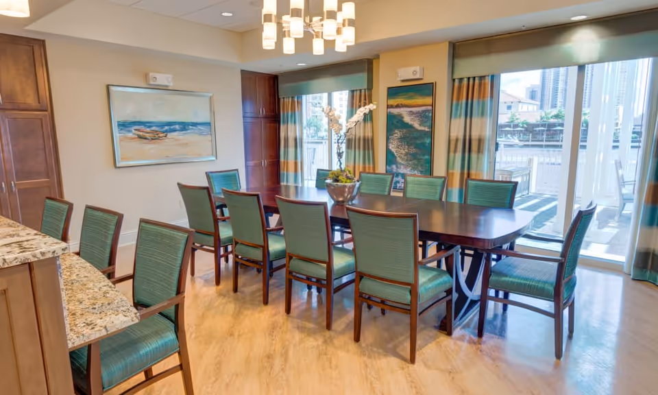 A bright dining room with a long wooden table surrounded by ten green upholstered chairs. The room features large windows with striped curtains, allowing natural light to fill the space. There are two paintings on the walls, one depicting a boat on the beach and the other a coastal scene. A modern chandelier hangs from the ceiling, and a granite countertop with additional chairs is visible on the left side.
