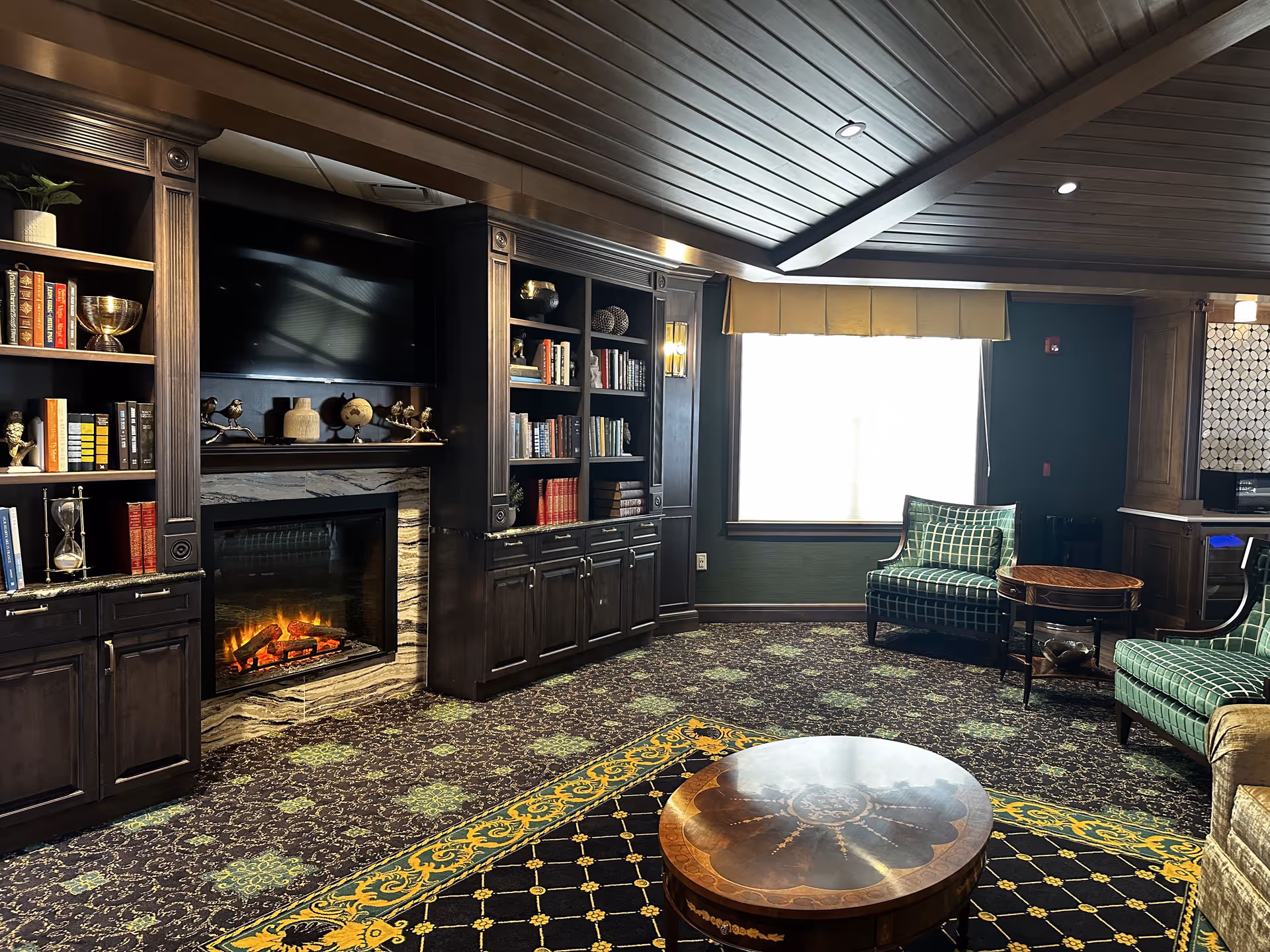 A cozy living room area with dark wood built-in bookshelves and cabinets surrounding a fireplace with a TV mounted above it. The room features patterned carpet with green and yellow accents, two green checkered armchairs, a round wooden coffee table, and a window with a beige valance letting in natural light. The ceiling has dark wood paneling with recessed lighting.
