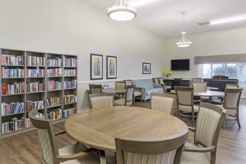 A well-lit common area with multiple round wooden tables surrounded by chairs. There is a bookshelf filled with books on the left side, a light blue couch along the back wall, framed artwork on the walls, a flat-screen TV mounted on the wall, and large windows with blinds letting in natural light.