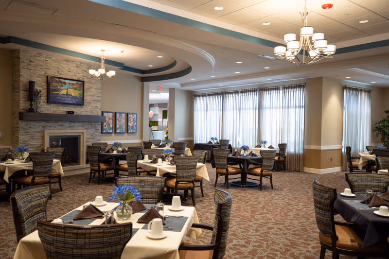 A spacious dining room with multiple tables covered in beige and black tablecloths, each set with white cups, brown napkins, and small flower vases. The room features large windows with sheer curtains, a stone fireplace with artwork above it, and elegant chandeliers hanging from the ceiling.