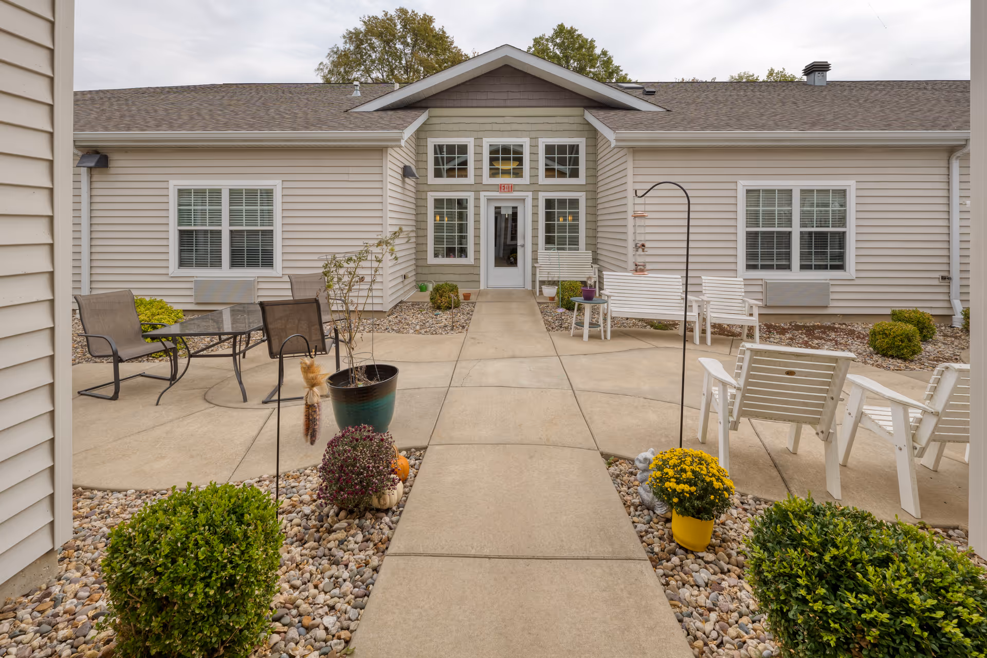 Outdoor patio area at a senior living facility with concrete walkways, potted plants, bushes, and outdoor seating including white chairs and a glass table with chairs. The building exterior is beige with white trim and multiple windows.