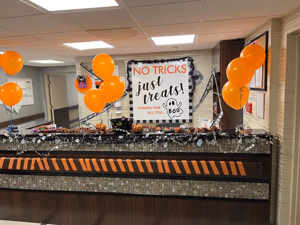 Reception desk decorated for Halloween with orange balloons, black and orange striped ribbon, small pumpkin decorations, and a sign that reads 'No tricks just treats! Thanks for all you... Boo' in a senior living facility.