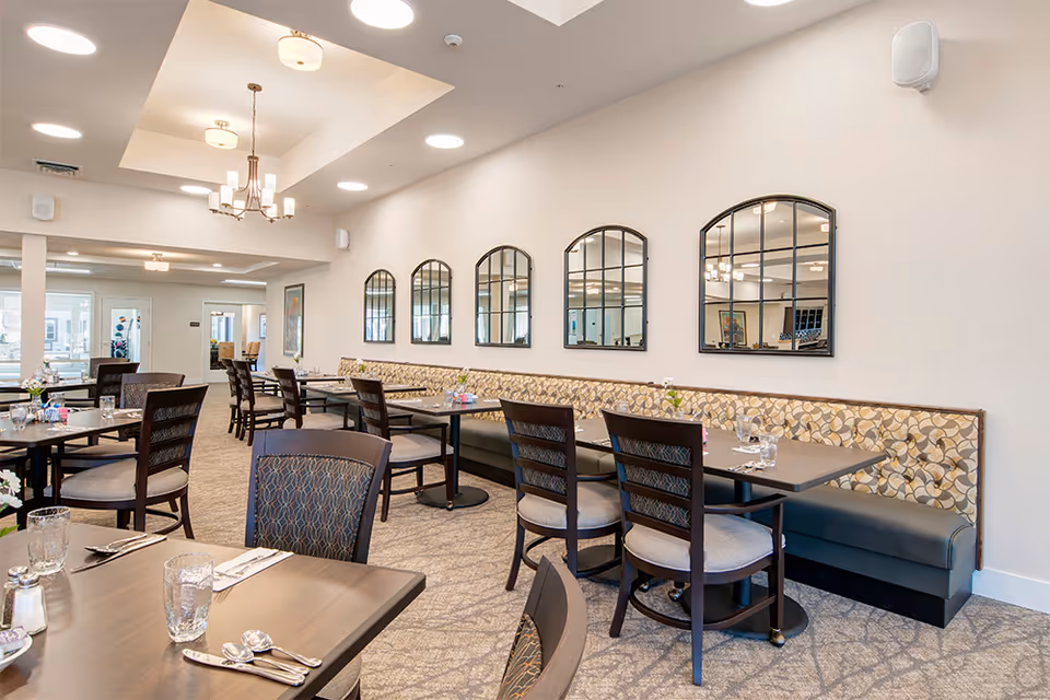 A bright and spacious dining room in a senior living community featuring multiple tables set with glasses, silverware, and napkins. The room has patterned cushioned bench seating along one wall with five arched mirrors above it. The ceiling has recessed lighting and a chandelier, and the floor is carpeted.