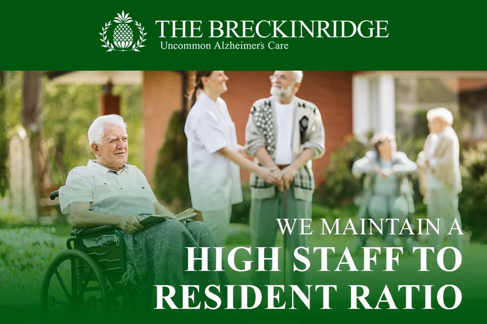 An elderly man in a wheelchair holding a book outdoors with a caregiver and other elderly people in the background, in a garden or courtyard setting at The Breckinridge Memory Care facility.