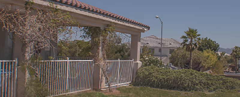 Exterior view of a residential building with a covered porch supported by columns, a metal railing, and surrounding greenery including bushes and trees under a clear blue sky.