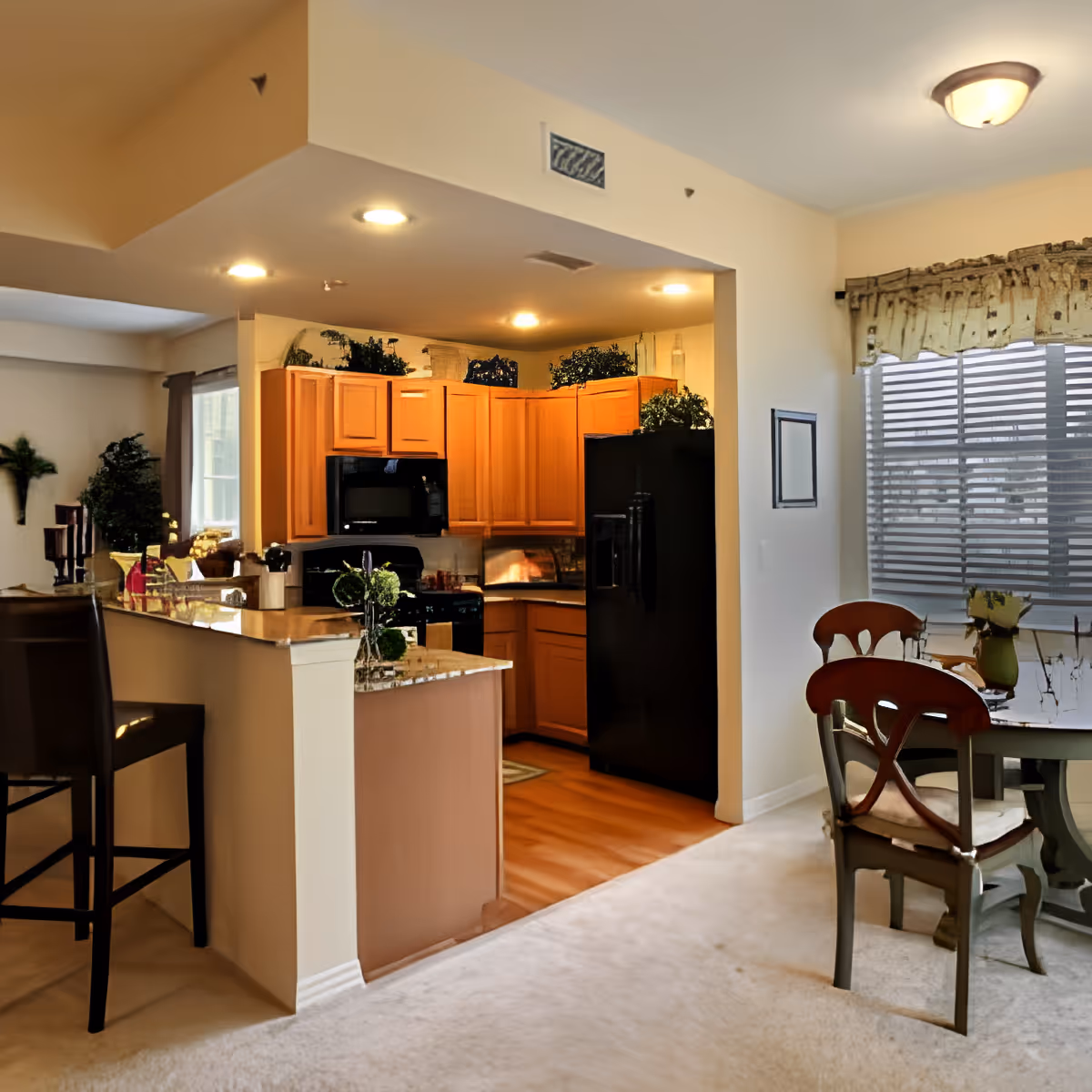 Interior view of a senior living facility showing a kitchen with wooden cabinets, black appliances including a refrigerator and microwave, and a granite countertop with bar stools. Adjacent to the kitchen is a dining area with a round table and chairs near a window with blinds and a valance. The space is well-lit with ceiling lights and decorated with plants and small decorative items.