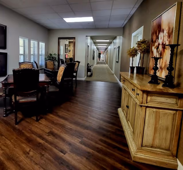Interior view of a senior living facility showing a dining area with a wooden table and chairs, a sideboard with decorations, and a long corridor leading down the hall.