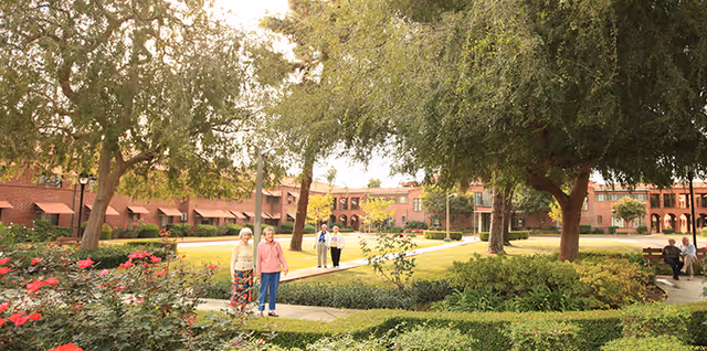 A landscaped outdoor garden area at Claremont Manor Retirement Community with green lawns, trees, bushes, and blooming flowers. Several elderly people are walking and sitting on benches along paved pathways. The background shows a large brick building with arched windows and awnings.