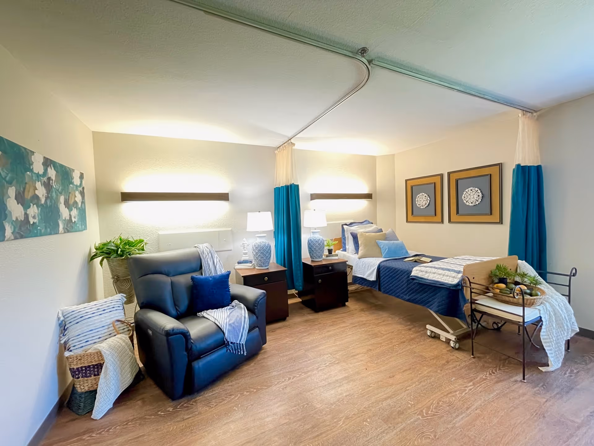 A well-lit senior living bedroom featuring a hospital-style bed with blue and beige bedding, two bedside tables with matching lamps, a comfortable dark blue recliner chair with a blue pillow and a throw blanket, a basket with blankets, decorative wall art, and a small table with a basket of fruit and plants. The room has wood flooring and light-colored walls with teal curtains around the bed.
