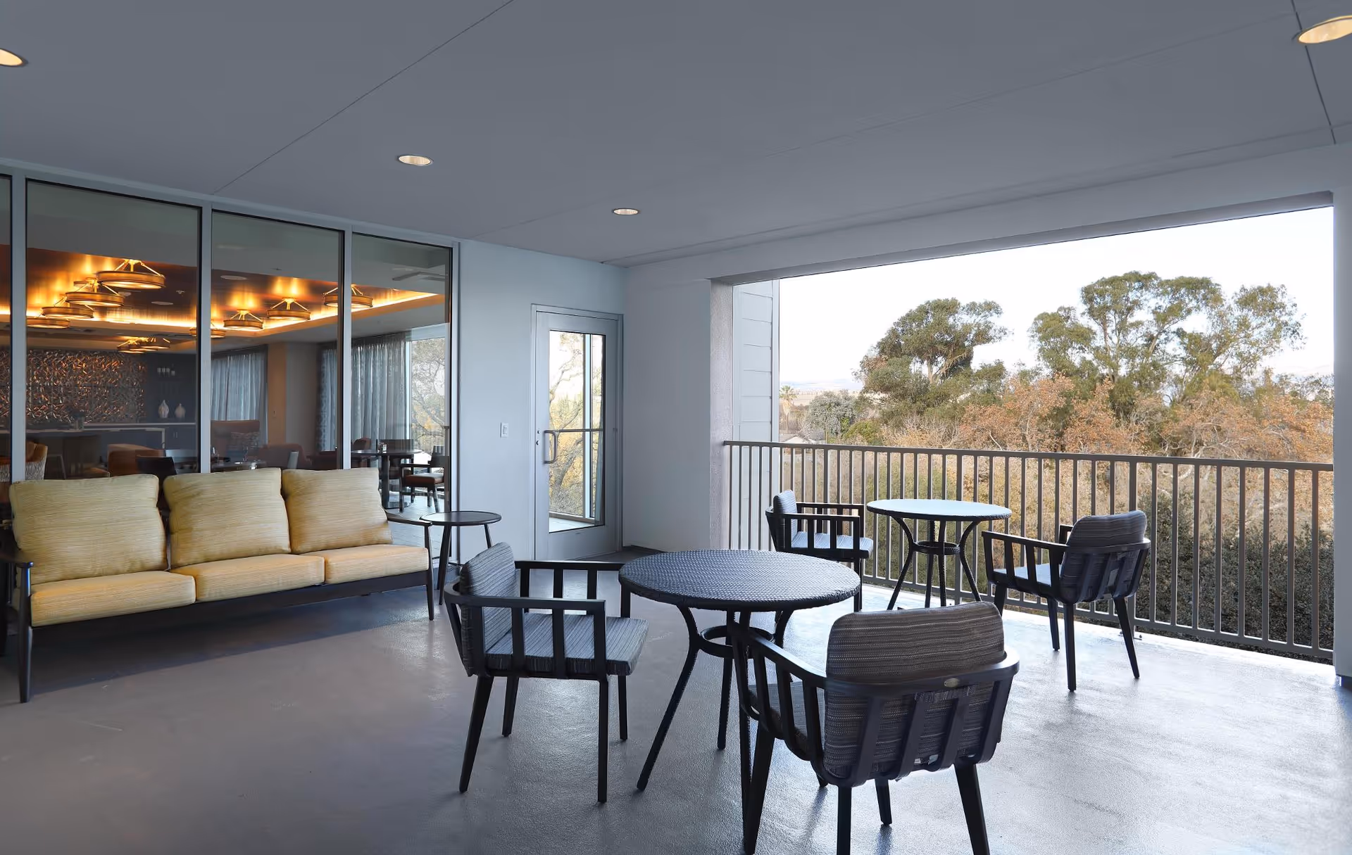 Covered outdoor patio area with a yellow cushioned sofa, black round tables, and black chairs with cushions. The patio overlooks trees and has a railing along the edge. Inside, through glass doors, a dining area with tables and chairs is visible.