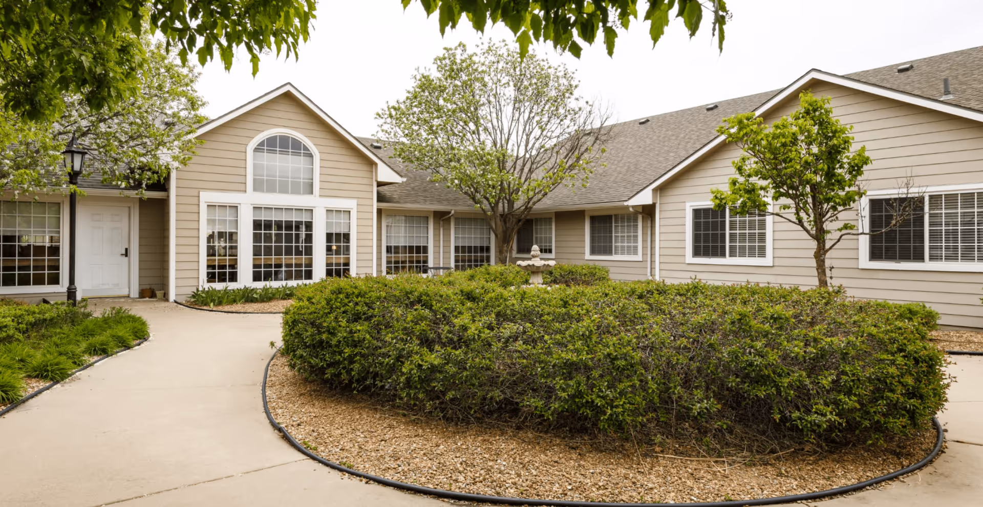 Front exterior of a single-story senior living building with large windows, shrubs, a circular walkway and a central fountain.