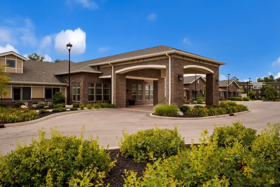 Exterior view of a senior living facility building with a covered entrance, brick walls, and a driveway surrounded by landscaped greenery under a blue sky with some clouds.