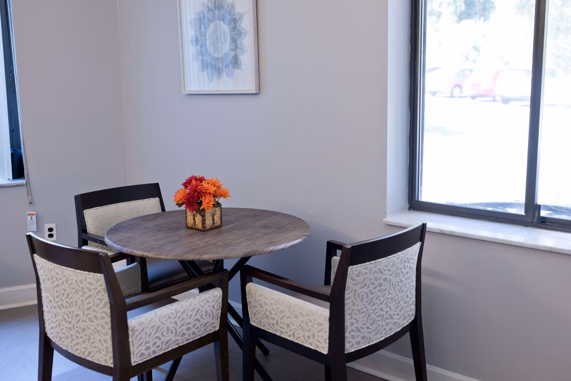 A small round wooden table with a decorative flower arrangement in a square vase on top, surrounded by four cushioned chairs with patterned upholstery. The room has light-colored walls, a window letting in natural light, and a framed artwork hanging on the wall.
