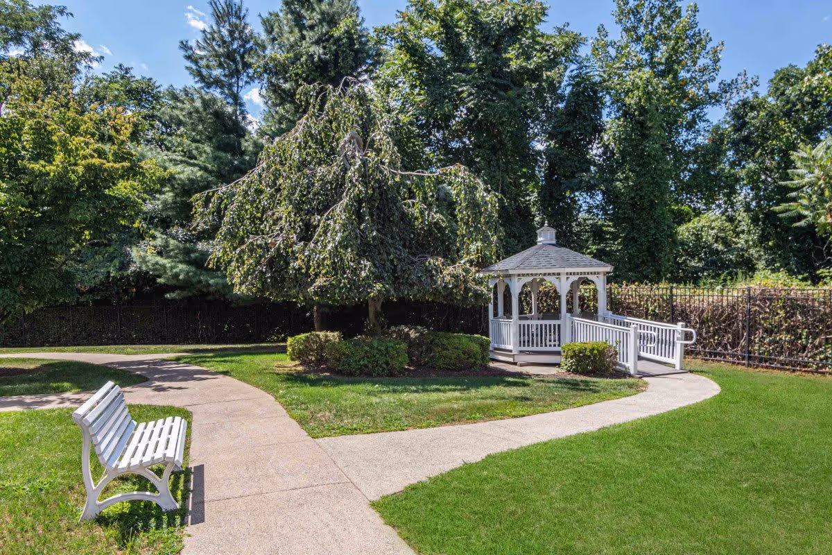 A peaceful outdoor garden area with a white wooden bench on a concrete pathway, a small white gazebo with a ramp, surrounded by green grass, bushes, and tall trees under a blue sky with some clouds.