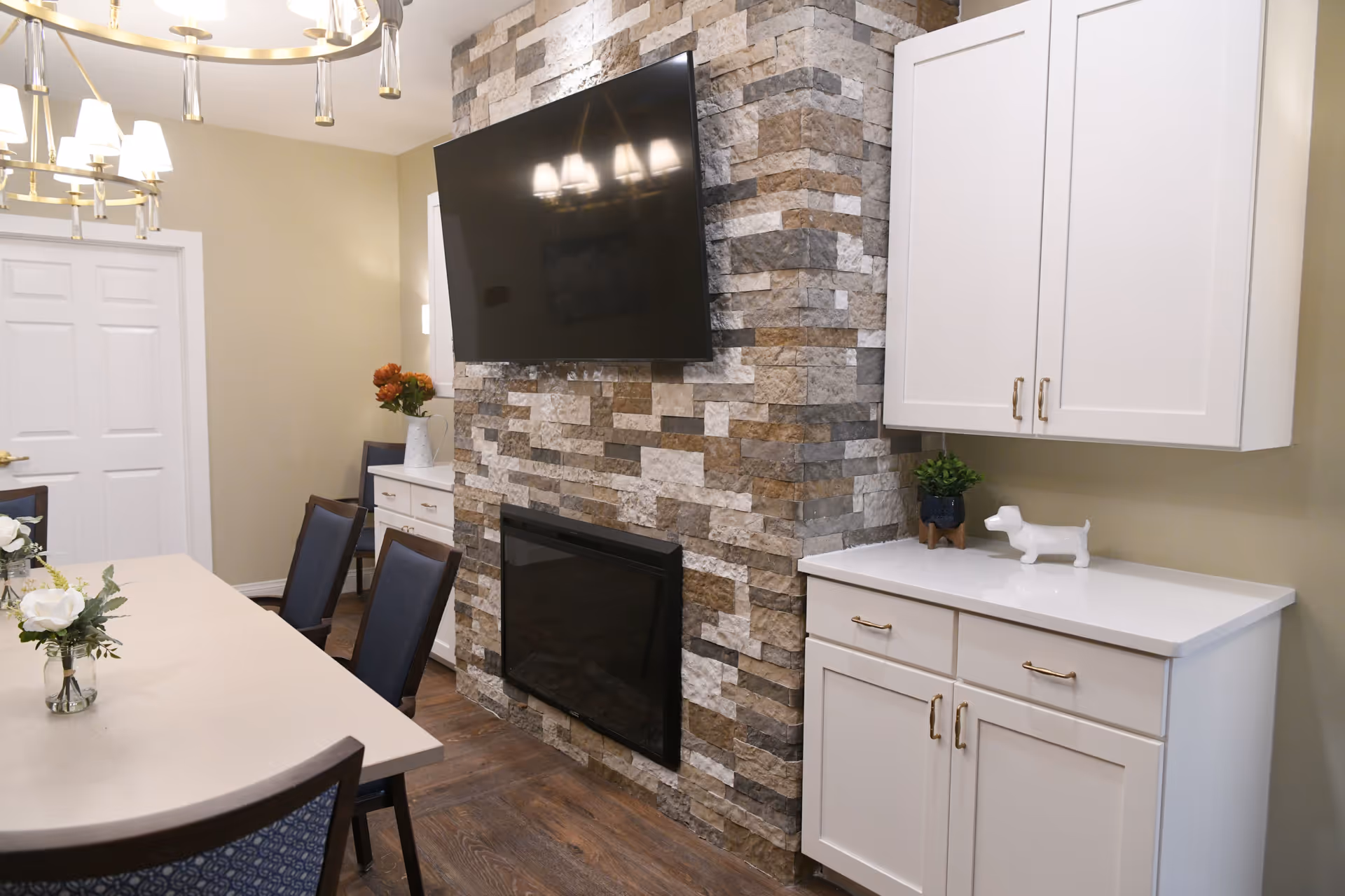 A dining area with a long table and chairs, a stone accent wall with a mounted flat-screen TV above a fireplace, white cabinets with gold handles, and decorative items including flowers and a small dog figurine.