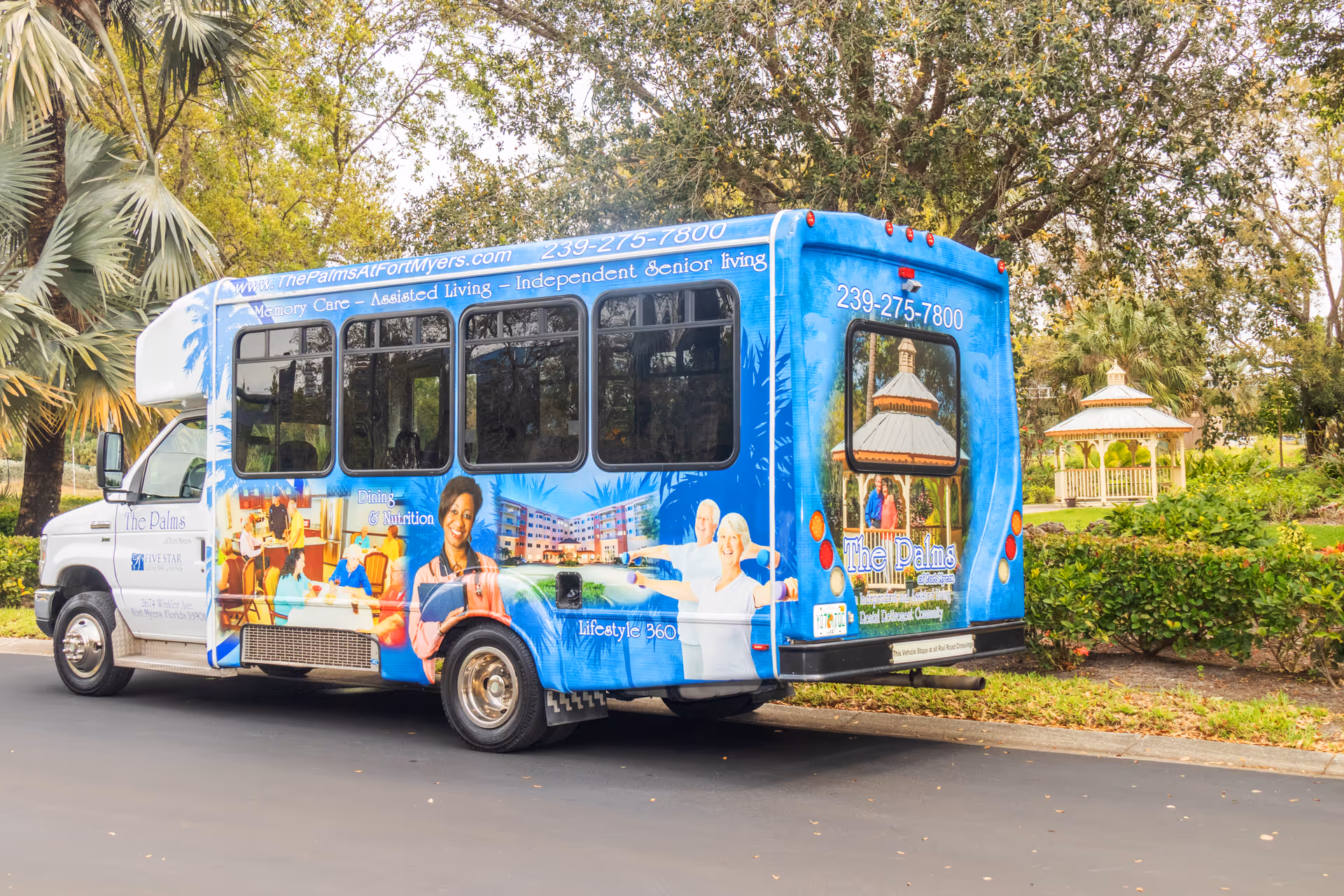 A blue shuttle bus parked on a road near greenery and a gazebo. The bus is branded with The Palms of Fort Myers senior living facility information, including images of smiling seniors, a building, and text about memory care, assisted living, and independent senior living.