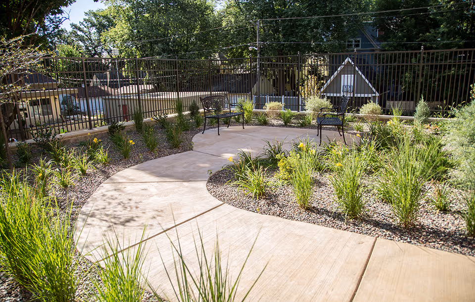 Curved paved garden walkway with benches, ornamental grasses, and a metal fence enclosing a landscaped courtyard.