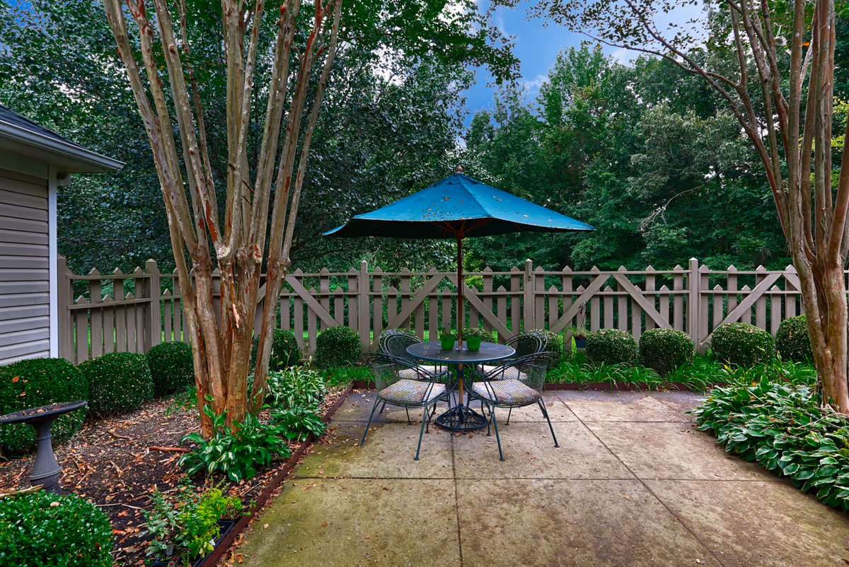 Outdoor patio area with a round metal table and four chairs, shaded by a large blue umbrella. The patio is surrounded by neatly trimmed bushes, trees, and a wooden fence, with a birdbath visible on the left side.