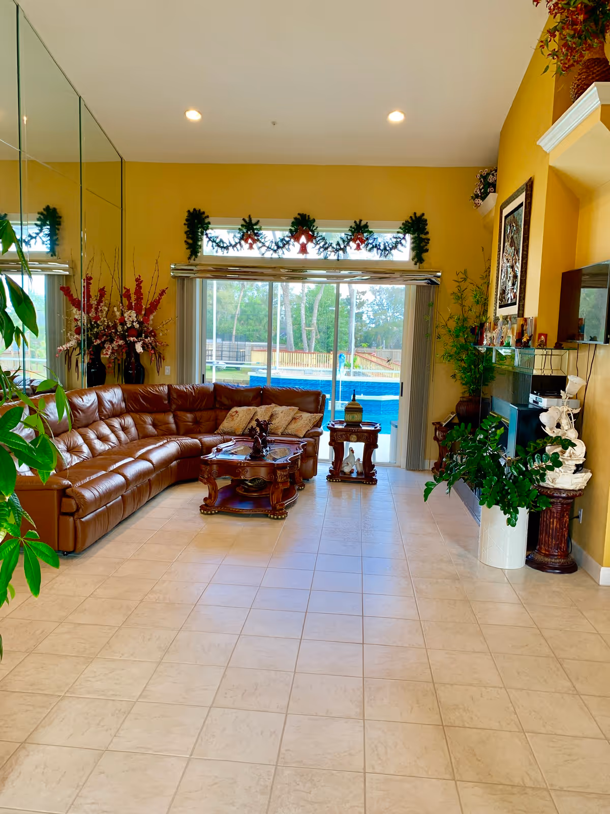 A spacious living room with a large brown leather sectional sofa, a wooden coffee table, and a side table. The room has yellow walls and a tiled floor. There is a large sliding glass door decorated with garlands and red bows, leading to an outdoor pool area. The room also features various plants, a fireplace with decorative items, and a large mirror on one wall.