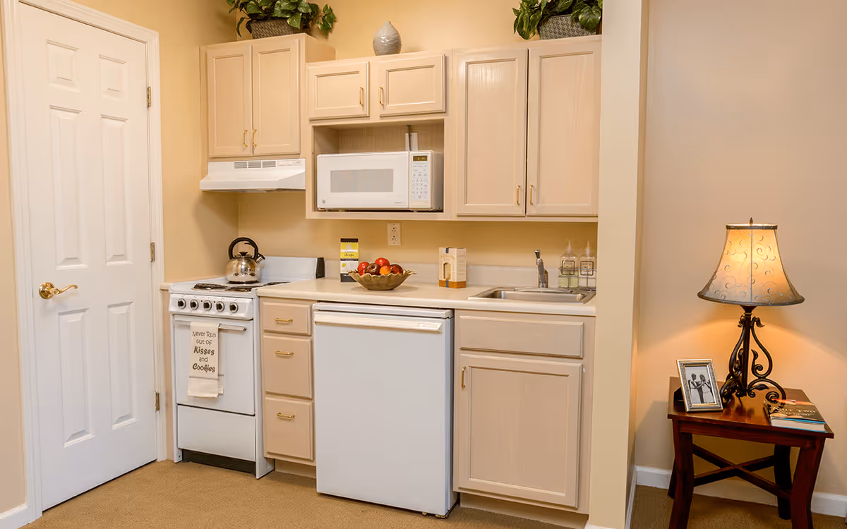A small kitchen area with light wood cabinets, a white microwave mounted above a white stove with a kettle on it, a white dishwasher, and a sink with soap dispensers. To the right, there is a wooden side table with a decorative lamp, a framed photo, and a book. The walls are painted beige and the floor is carpeted.