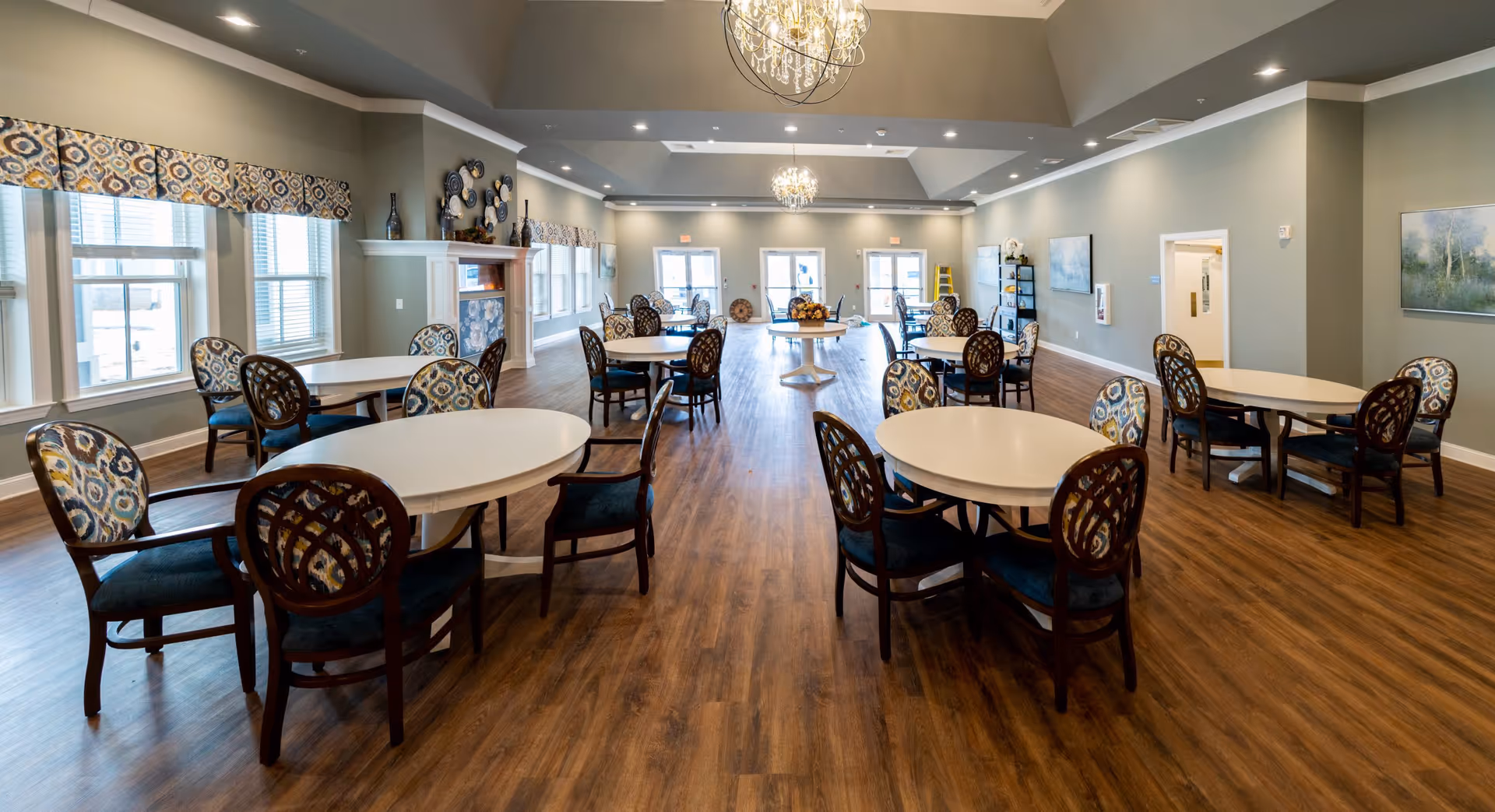A spacious dining room with multiple round tables and chairs featuring patterned upholstery. The room has large windows with matching valances, wooden flooring, and two chandeliers hanging from a high ceiling. There is a fireplace on the left wall and artwork on the right wall.