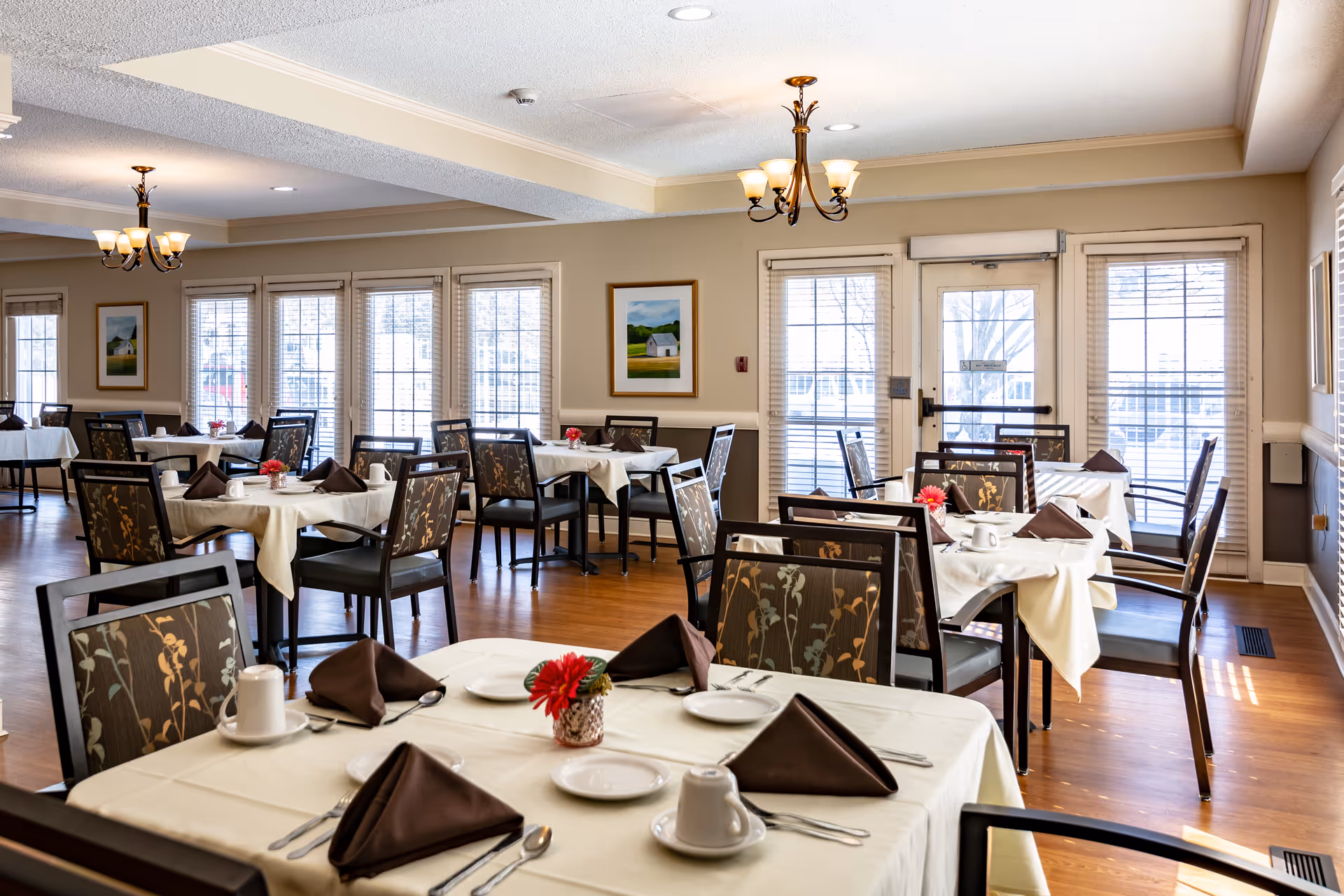 Well-lit dining room with neatly set tables and chairs, floral centerpieces, and large windows.