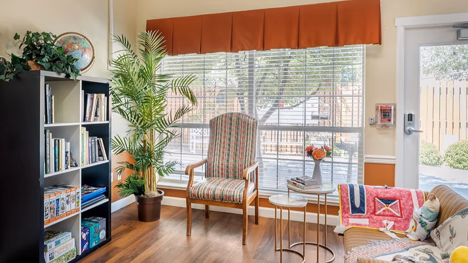 A cozy sitting area with a wooden floor, featuring a plaid upholstered armchair, a small round side table with a vase of flowers and books, a brown couch with colorful blankets and pillows, a black and white bookshelf filled with books and games, a potted plant, and a large window with blinds letting in natural light. A door with a keypad lock and a fire alarm is visible on the right.