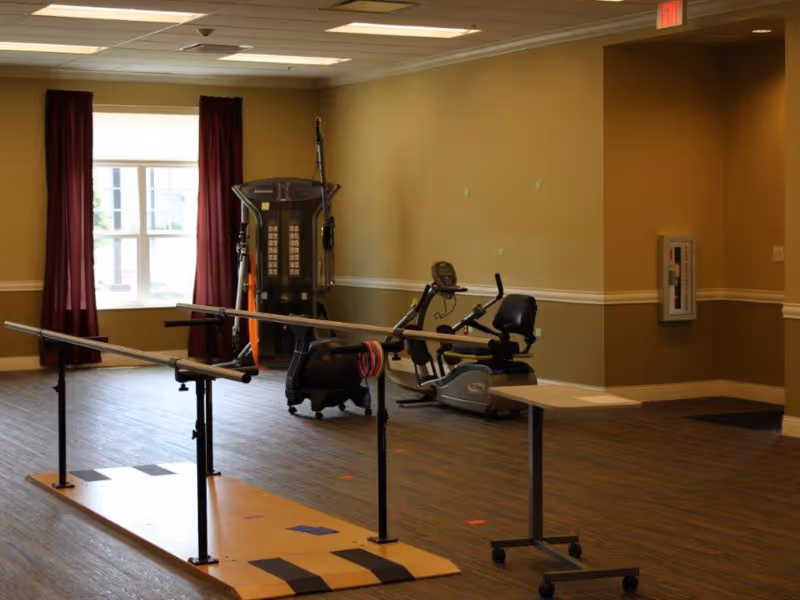 An indoor rehabilitation or exercise room with parallel bars for walking practice, a recumbent exercise bike, and other fitness equipment. The room has wood flooring, beige walls, a window with maroon curtains, and a small table on wheels.