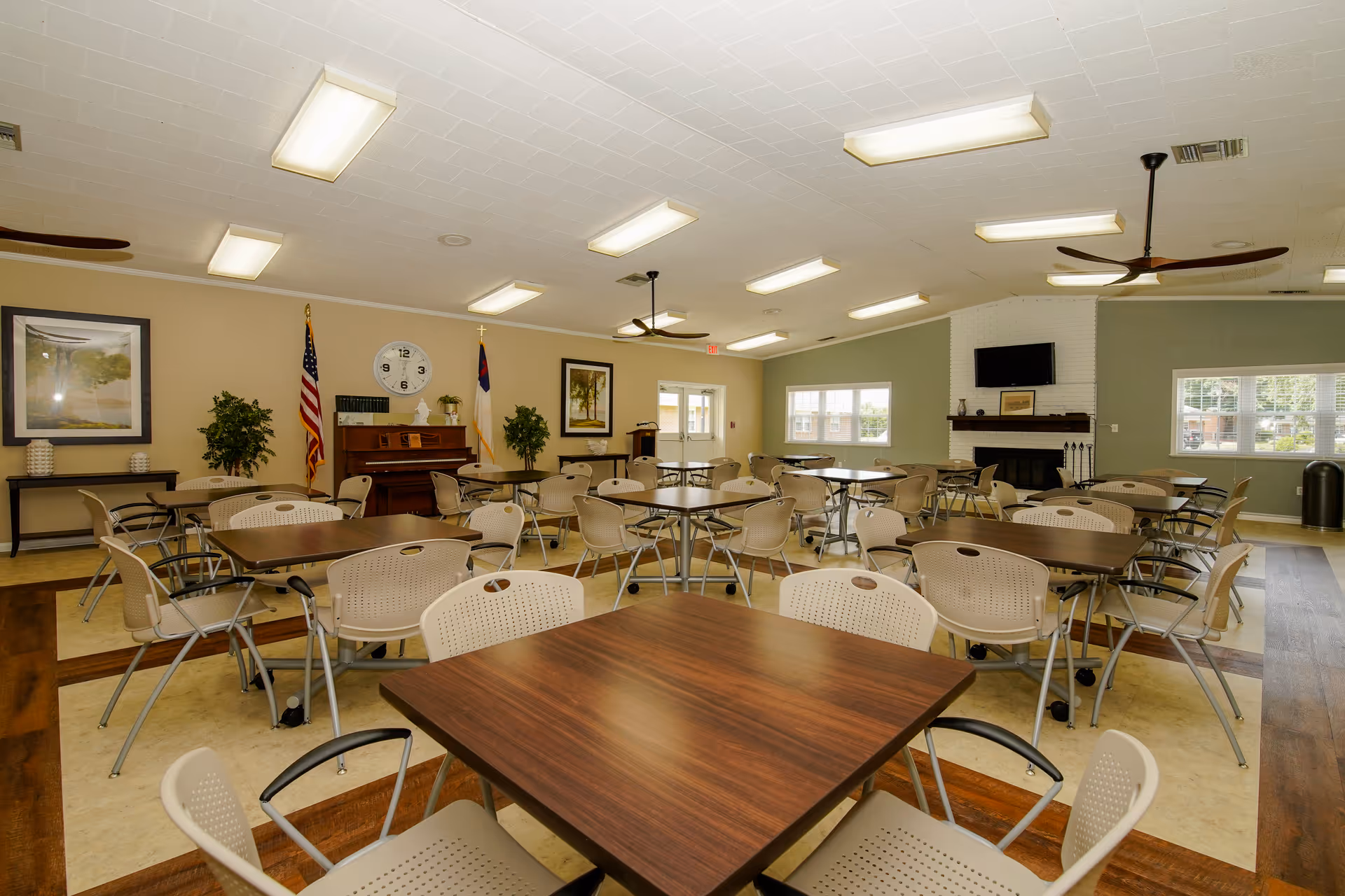 A spacious senior living facility common room with multiple square wooden tables and beige chairs arranged neatly. The room features a piano against the wall with an American flag and a Christian flag beside it, a large wall clock, framed artwork, two ceiling fans, and a fireplace with a mounted TV above it. Large windows allow natural light to enter the room.